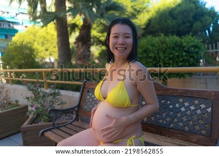 outdoors summer holidays portrait of young happy and attractive Asian Japanese woman pregnant at resort swimming pool enjoying carefree proud of her belly in maternity concept