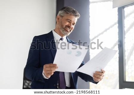 Happy Middle Aged Entrepreneur Reading Business Papers At Office, Smiling Mature Businessman In Suit Holding Documents, Checking Company Growth Rates While Standing At Workplace, Copy Space