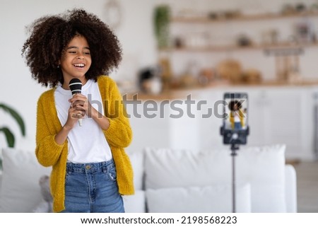 Positive pretty little black girl in casual singing and recording video, broadcasting from home, using microphone and modern mobile phone on tripod, sharing video with her friends, copy space Royalty-Free Stock Photo #2198568223