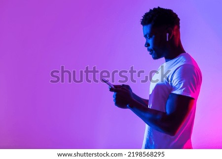 Side view of young man in earphones listening to music, using new mobile app on smartphone in neon light, free space. Cheerful African American guy enjoying new playlist or audio book
