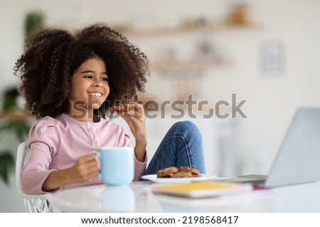 Cheerful girl with bushy hair having break while studying at home, sitting at desk, eating cookies, drinking juice or cocoa, looking at computer screen and smiling, watching cartoons, copy space