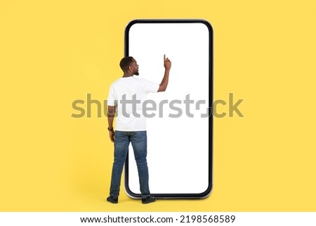Back View Of Black Man Using Huge Phone With Empty Screen Standing On Yellow Studio Background. Guy Using Mobile Application And Browsing Internet On Big Smartphone. Mockup
