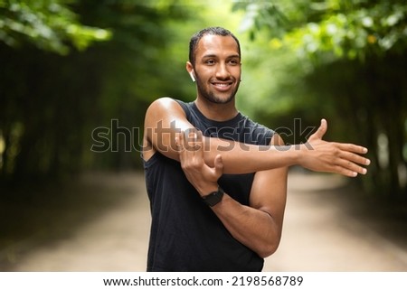 Closeup photo of cheerful sporty handsome young black guy having workout outdoors, stretching arms before jogging, using earbuds, listening to music while training, looking at copy space