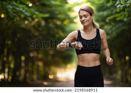 Athletic blonde woman looking at fitness bracelet while running alone outdoors, smiling middle aged woman in sportswear checking tracker on her wrist, exercising at public park, copy space