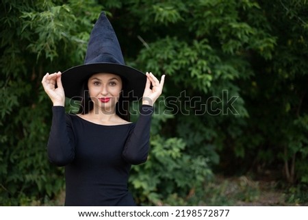 Halloween. A young beautiful woman in a witch's hat and a black dress in the park. Space for text.High quality photo