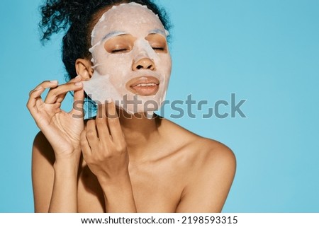 African American woman applies cosmetic tissue facial mask for hydrating and moisturizing and glowing face skin, on blue background. Skincare concept and daily self-care routine, high quality