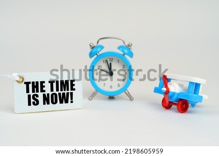 On a white surface there is a toy plane, an alarm clock and a sign with the inscription - The Time is Now. Business concept.