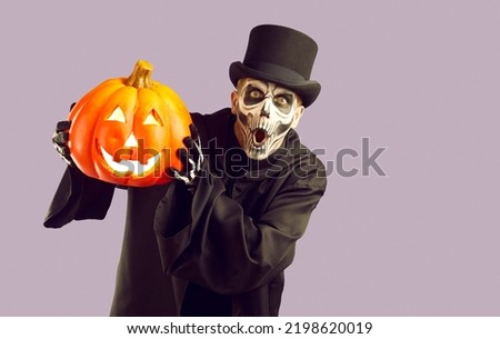 Man in Halloween costume. Skeleton in black cloak and top hat standing isolated on light purple background, holding orange jack o lantern and looking at camera with funny surprised face expression Royalty-Free Stock Photo #2198620019