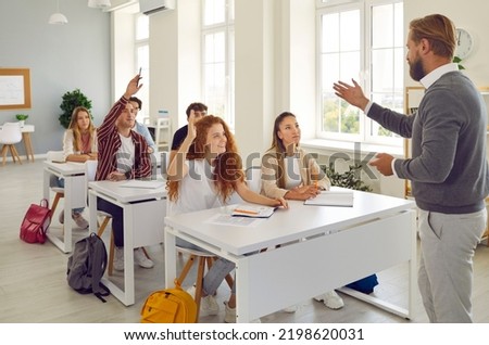 Learning process. Active college students raise their hands in class to answer teacher's questions. College or high school students sit at class desks and interact with male teacher. Royalty-Free Stock Photo #2198620031