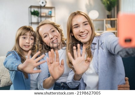 Attractive beautiful two people two young lady girls sisters and their mother using mobile phone taking photo, picture or having video call while sitting at home on sofa, waving hands.