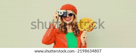 Autumn portrait of happy smiling young woman with film camera and yellow maple leaves wearing red french beret on gray background