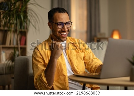 Joyful millennial black man with laptop celebrating online win or work success at home. Excited young African American guy enjoying big sale in web store, gambling on internet Royalty-Free Stock Photo #2198668039