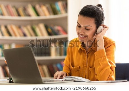 Knowledge concept. Mixed race woman wearing and touching wireless headphones, reading paper book while sitting in library with laptop, studying online