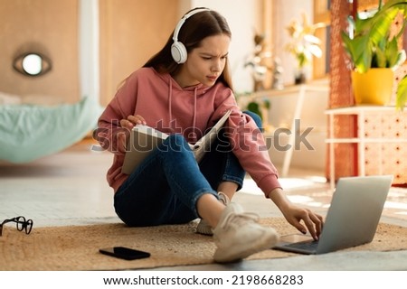 Concentrated teenage girl reading book in front of laptop, getting ready for exam, sitting on floor at home. Focused female student having remote lesson on pc Royalty-Free Stock Photo #2198668283