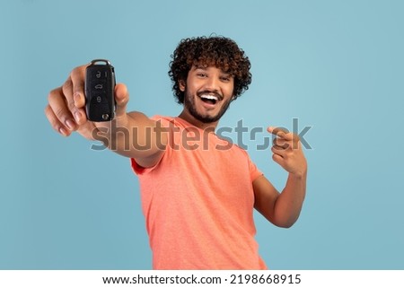 Cheerful happy excited vivid driver young bearded Indian man holding in hand new car key and pointing at it, isolated on blue background, studio shot, copy space. Driving, car leasing concept