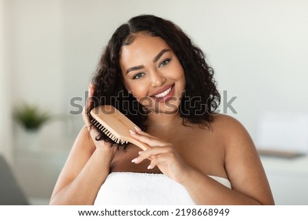 Happy chubby black lady making hair care, treatment and spa day at leisure time, combing curly healthy hair with comb in light bathroom and smiling at camera