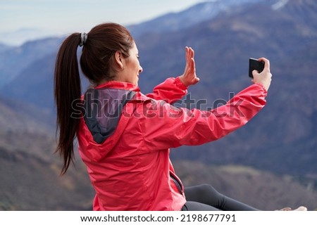 young woman with red hair on her side waving in a video call, wearing a red jacket, sitting on a rock in the middle of the Andes Mountains of Chile
