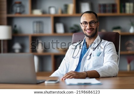 Handsome middl eastern young doctor sitting at worktable in front of laptop, posing at modern clinic, wearing eyeglasses, smiling at camera, copy space. Healthcare concept