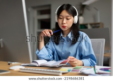 Korean Lady Designer Working With Color Samples Sitting Near Computer Wearing Headphones At Workplace In Modern Office. Young Graphic Designer Choosing Palette. Creative Career And Freelance