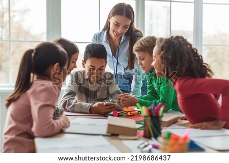 Multiethnic School Kids And Teacher Using Smartphone And Educational Application Learning Sitting At Desk In Modern Classroom At School. Education And Gadgets Concept. Selective Focus