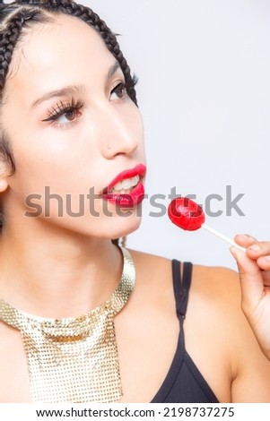 Head and shoulders portrait of young Latina woman with red lipstick, holding a red candy pop as it approaches her lips.