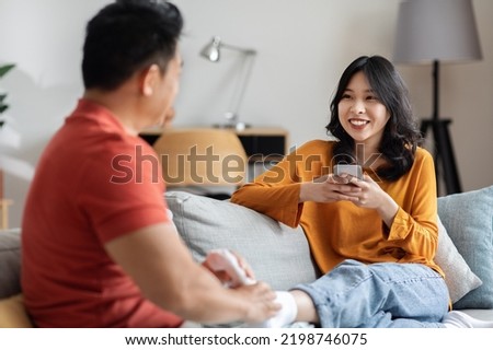 Loving asian husband massaging his smiling wife feet, happy chinese young woman relaxing on couch at home, using smartphone, having conversation with boyfriend. Love, affection, relationship