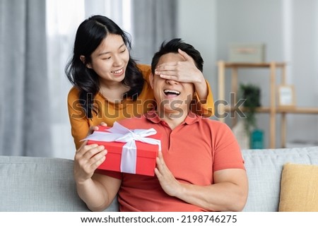 Loving asian woman closing eyes from behind and giving gift box her excited boyfriend, making great surprise for St. Valentines Day or birthday, home interior, free space. Anniversary celebration