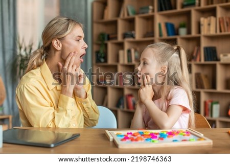 Professional woman speech therapist studying together with pretty little girl, learning practice pronunciation exercises with child at office, touching faces and grimacing, sitting at desk Royalty-Free Stock Photo #2198746363