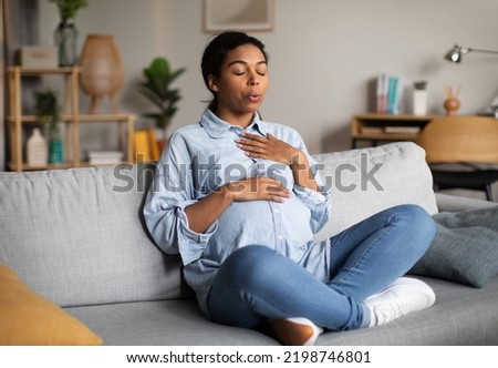Pregnancy Yoga. Pregnant African American Woman Doing Breathing Exercise With Eyes Closed Relaxing Sitting On Couch At Home. Relaxation And Childbirth Healthcare Concept Royalty-Free Stock Photo #2198746801