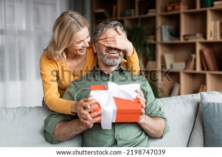Loving Wife Surprising Middle Aged Husband With Present, Covering His Eyes And Giving Gift Box, Caring Woman Greeting Spouse With Birthday While They Resting Together In Living Room At Home