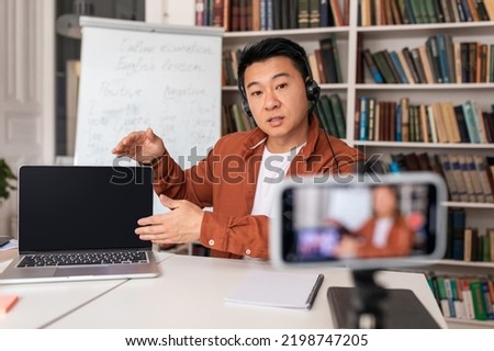 Asian Male Teacher Having Online Lecture Showing Laptop Computer With Empty Screen Filming Lesson On Smartphone In Modern Classroom, Wearing Headset. E-Teaching. Selective Focus