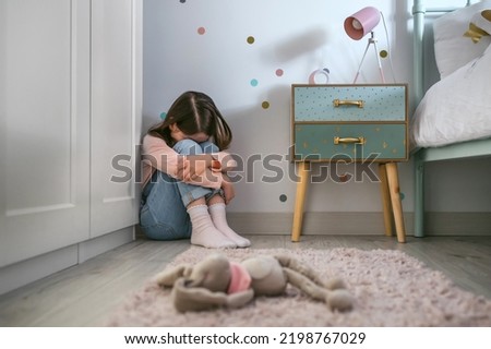 Unrecognizable sad little girl sitting on the floor of her bedroom with stuffed toy lying. Selective focus on girl in background Royalty-Free Stock Photo #2198767029