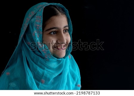 Little Muslim girl with beautiful eyes wearing blue hijab smiling with someone, black background, side view