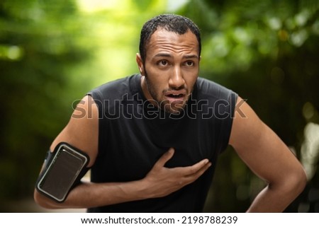 Handsome young black sportsman with heart pain touching his chest, jogging in the public park, using armband with smartphone, copy space. Sport and cardiovascular diseases concept Royalty-Free Stock Photo #2198788239