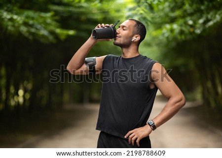 Sporty young black man drinking protein cocktail, training at public park, using wireless earbuds, listening to music while jogging by forest, copy space. Sports nutrition concept Royalty-Free Stock Photo #2198788609