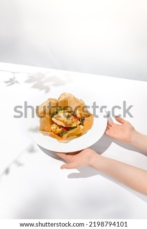 Female hand holding fish dish. Roasted fish in hand of person. Food menu with hand. Person eat baked fish in white plate with shadows. Trendy food menu. Take out food in elegant style