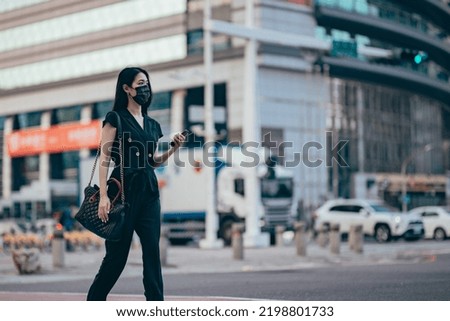  young Asian woman with protective face mask using smartphone on the go, against illuminated neon commercial signs in city street in downtown district at night.