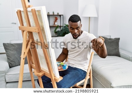 Young african man painting on canvas at home pointing down with fingers showing advertisement, surprised face and open mouth 