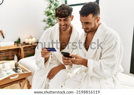 Two hispanic men couple holding credit card and smartphone sitting on massage table at beauty center