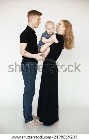 Portrait of young smiling beautiful family in dark clothes standing with little plump grey-eyed baby infant on white background. Mother holding child with hands. Family, relationship, having fun.