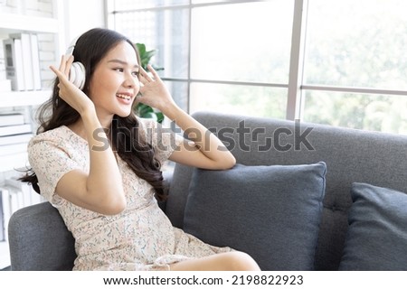 Relaxing casual asian  woman, casual cloth listening to music with headphone from streaming online playing music playlist application on smartphone on sofa couch at co working space. work life balance