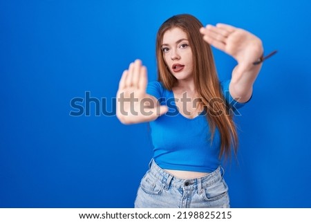 Redhead woman standing over blue background doing frame using hands palms and fingers, camera perspective 