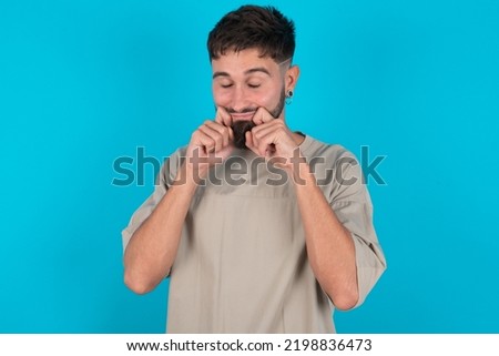 Pleased bearded caucasian man wearing casual T-shirt over blue background with closed eyes keeps hands near cheeks and smiles tenderly imagines something very pleasant