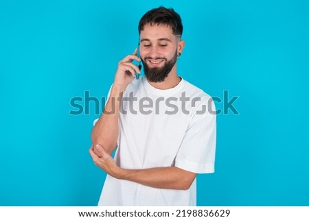 Portrait of a smiling bearded caucasian man wearing white T-shirt over blue background talking on mobile phone. Business, confidence and communication concept.