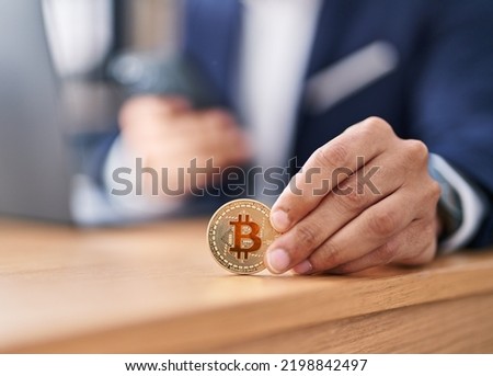 Young hispanic man business worker holding bitcoin using smartphone at office