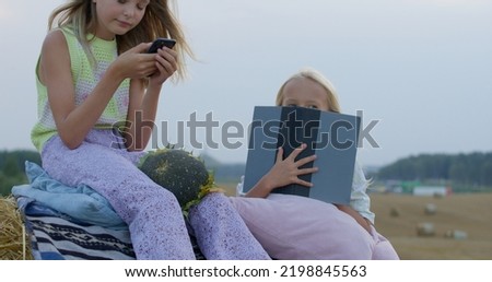 two girls eat sunflower seeds on mown rye in field. Landscape of straw bales against setting sun on background. Sisters watch video on smartphone and read book outdoors. Childhood. Country life