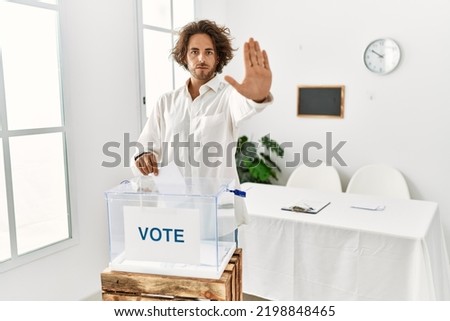 Young hispanic man voting putting envelop in ballot box doing stop sing with palm of the hand. warning expression with negative and serious gesture on the face. 