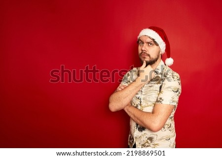 Puzzled confused young traveler tourist man in summer casual clothes christmas santa hat put hand on chin isolated on red studio background. Passenger traveling on holiday. Air flight journey concept