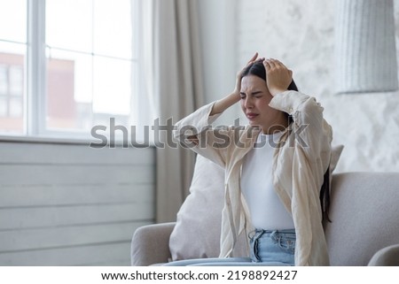Nervous breakdown, depression, panic attack. A young beautiful brunette woman sits on the sofa at home and holds her head with her hands, is nervous, feels pain, discomfort. Royalty-Free Stock Photo #2198892427