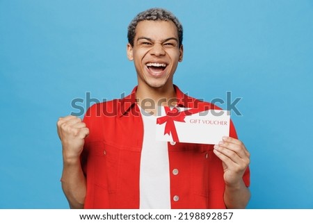 Young surprised happy man of African American ethnicity 20s he wear red shirt hold gift certificate coupon voucher card for store do winner gesture isolated on plain pastel light blue cyan background